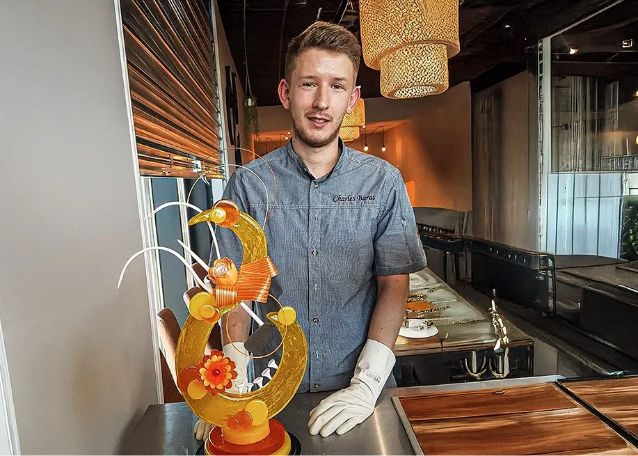 Charles Baras en veste de chef pâtissier à son nom, devant une pièce artistique en sucre
